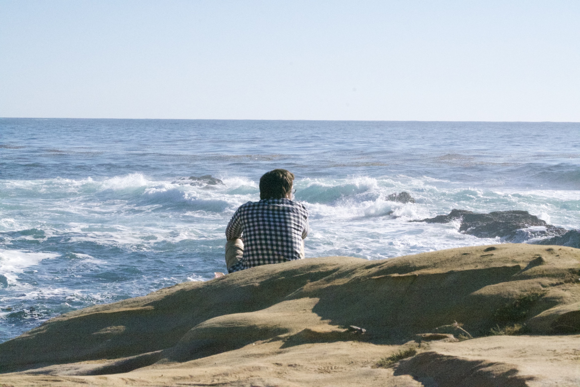 A man sits on rocky ground, looking at the ocean.