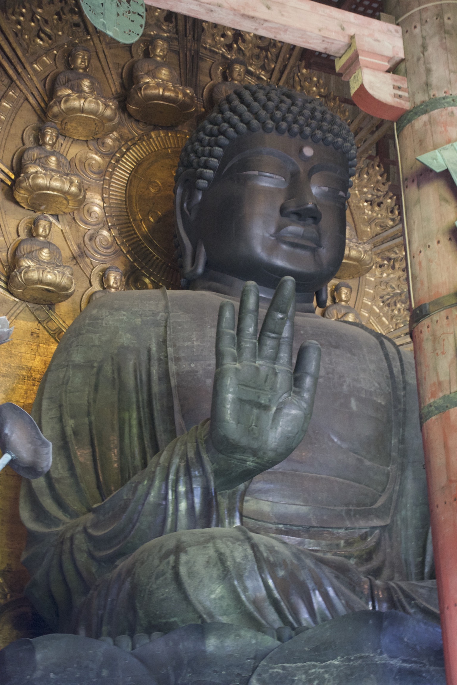 A bronze buddha with its right hand raised, seated before a golden backdrop of seated buddhas.