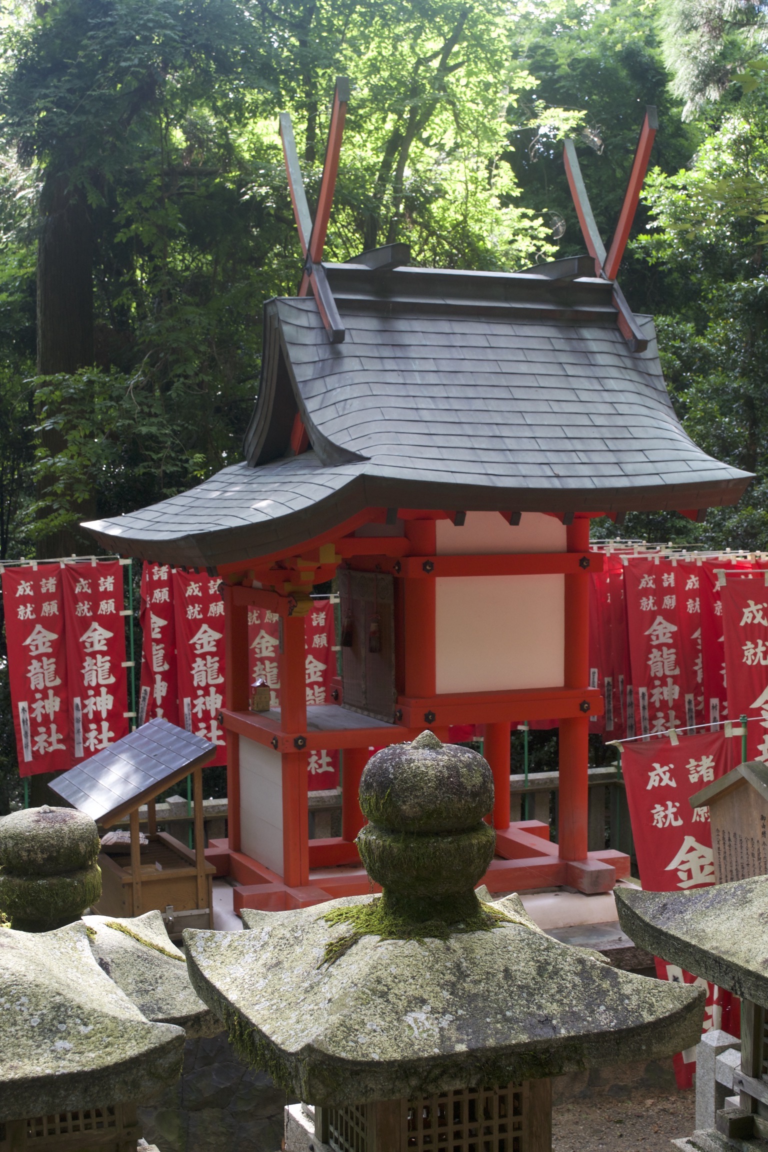 A white shrine with red trim and a slanted black roof sits before some mossy stone monuments.  Red banners with white lettering hang before the trees in the background.