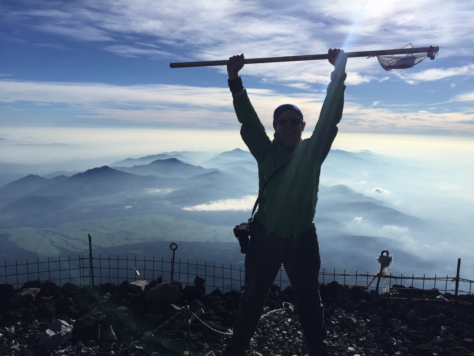 A man triumphantly holds his hiking stick above his head, atop a mountain.